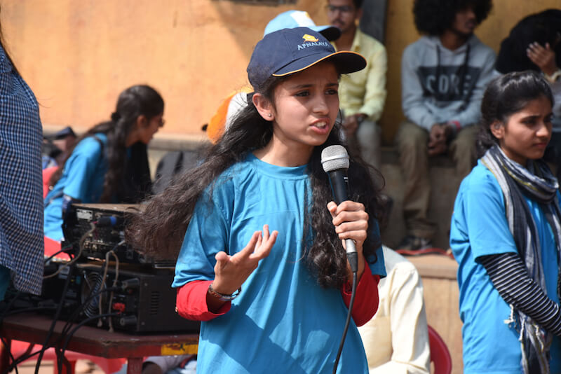 A young girl in a blue tunic and cap speaks passionately into a microphone at an outdoor event, with attentive onlookers in the background.