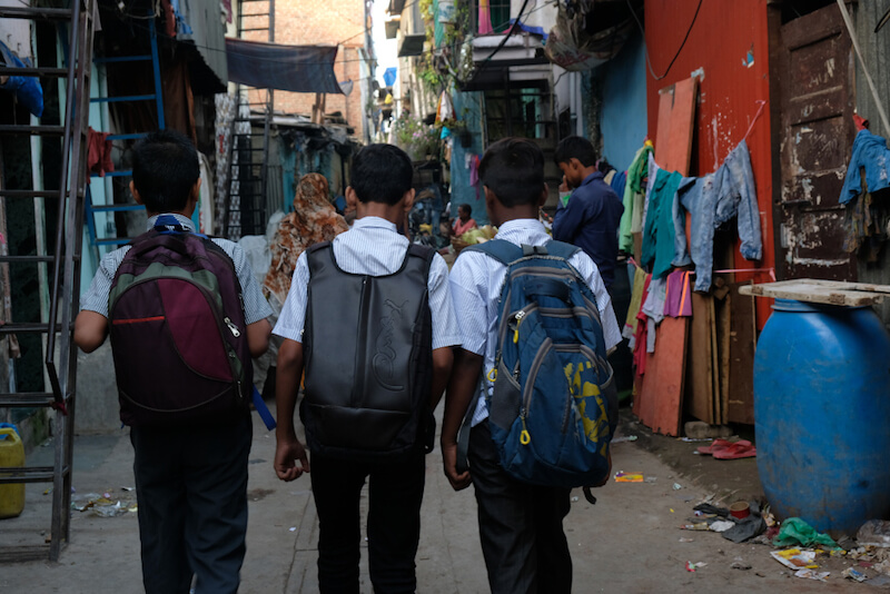 Three schoolboys with backpacks walk away from the camera down a narrow alley lined with houses and hanging clothes in an urban setting.
