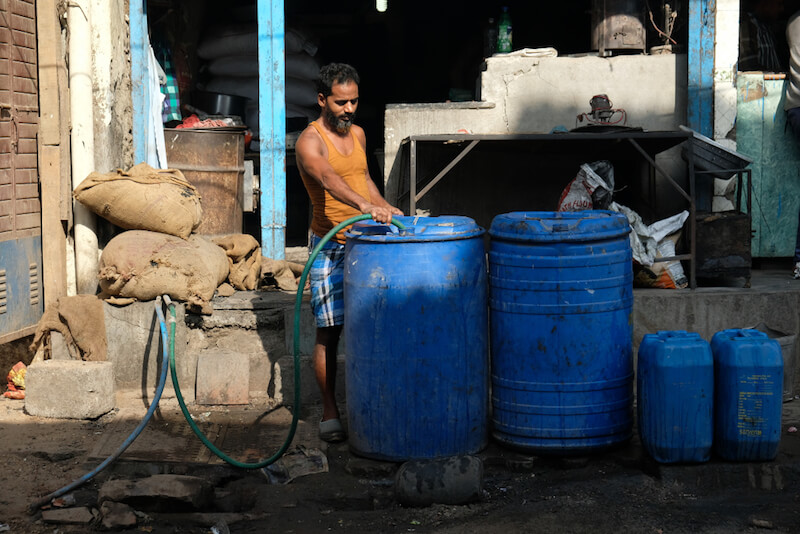 A man in a sleeveless shirt fills blue water drums with a hose, set against a backdrop of a cluttered street scene, suggesting a water distribution point.