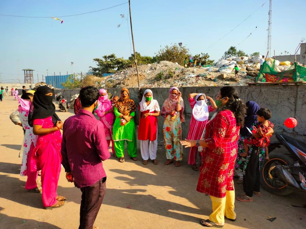 A vibrant group of people, mostly women in colorful attire, gathers on a sunny day in a street, indicative of a community event.