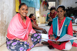 A smiling woman in a pink shawl and patterned skirt participates in a group learning session with other women in traditional Indian clothing, inside a simple, informal setting.